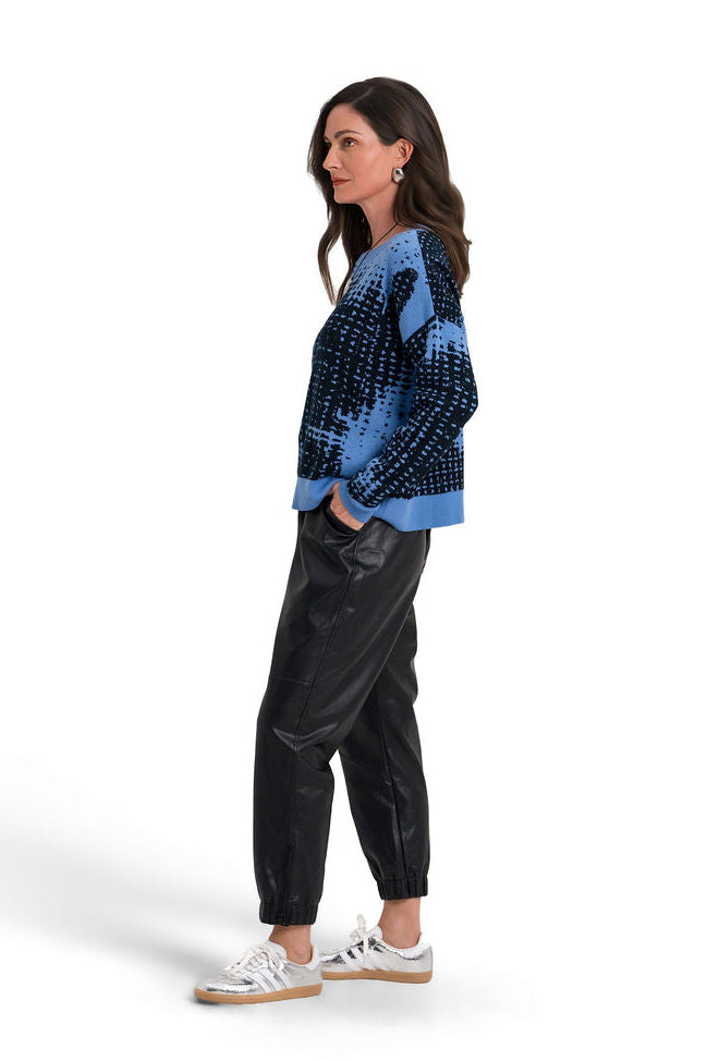 A woman stands in profile wearing a blue and black patterned pima cotton knit sweater, black pants, and silver sneakers against a plain white studio background.