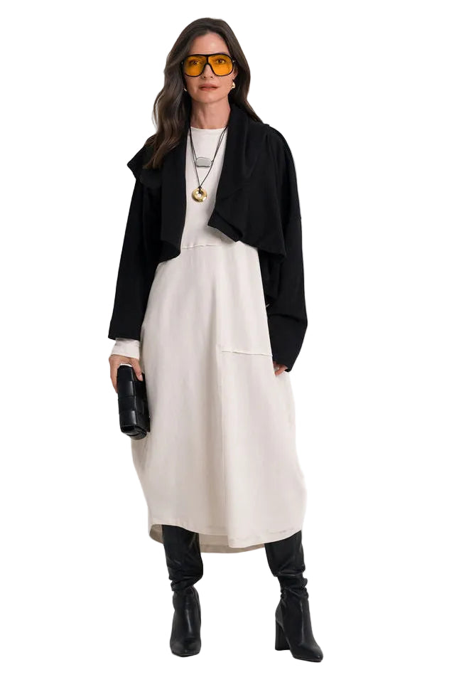 A model stands against a plain white studio background wearing a 100% cotton black cropped cardigan over a cream midi dress, with black knee-high boots, layered necklaces, and orange-tinted sunglasses.