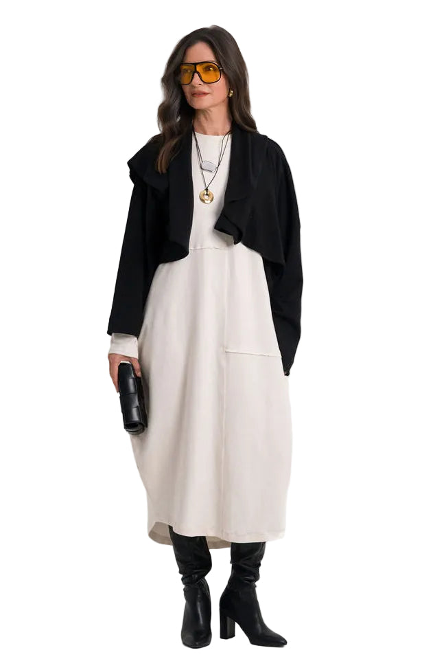 A woman modeling a 100% cotton black cropped cardigan over a cream-colored midi dress, styled with black heeled boots, gold-framed sunglasses, and a small black clutch, set against a plain white studio background.