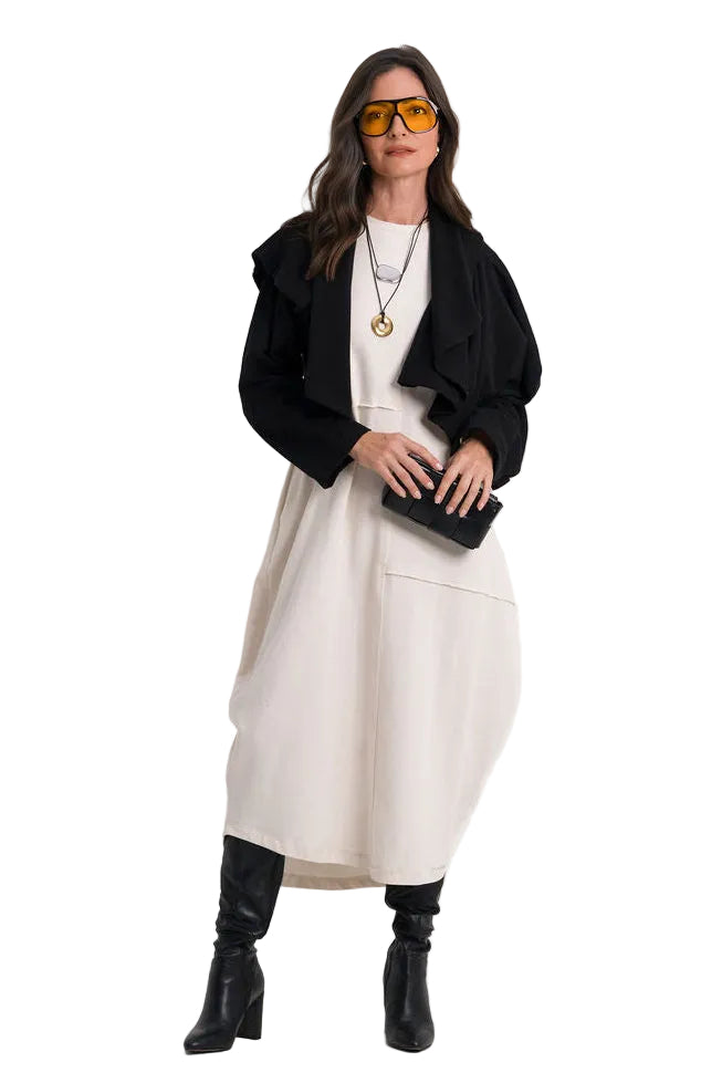 A woman modeling a 100% cotton black cropped cardigan over a cream-colored midi dress, styled with black knee-high boots, a black clutch, and yellow-tinted sunglasses against a plain white studio background.