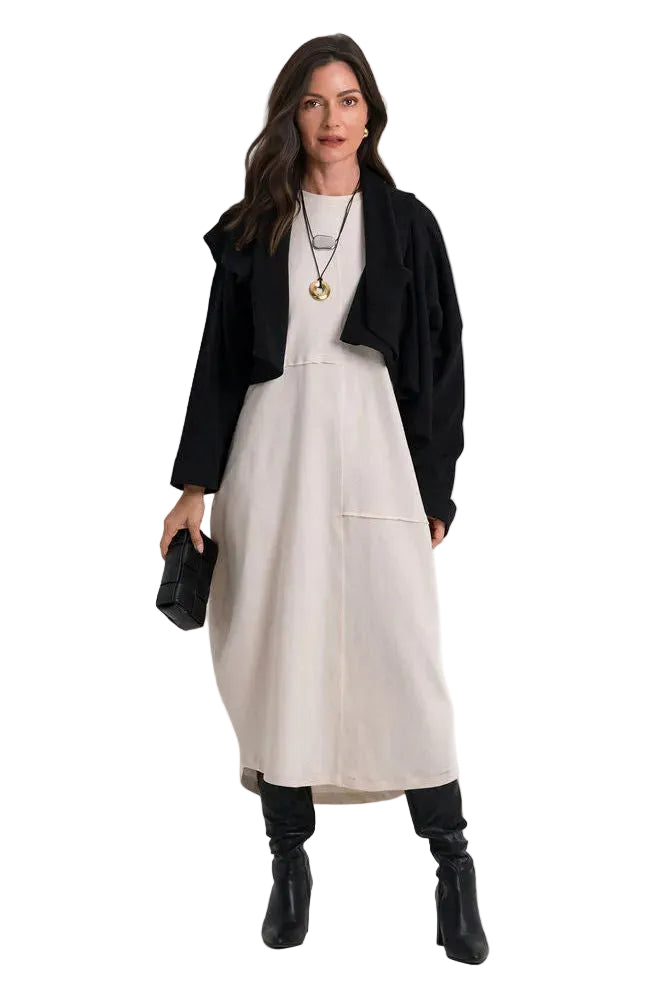 A woman modeling a 100% cotton black cropped jacket over a light beige midi dress, styled with black boots, a gold pendant necklace, and holding a small black clutch bag against a plain white studio background.