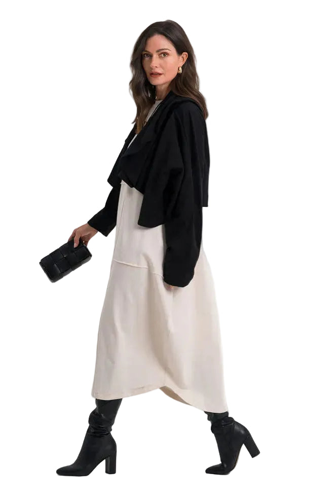 A woman models a 100% cotton black cropped cardigan layered over a cream-colored maxi dress, paired with black heeled ankle boots and holding a small black clutch, set against a plain white studio background.