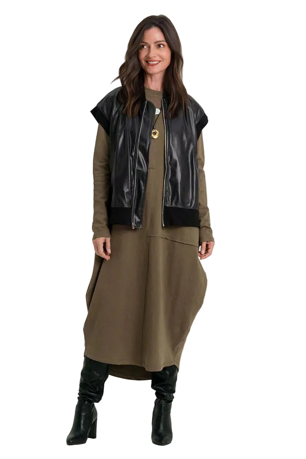 A woman modeling a 100% cotton long olive green dress with a shiny black faux-leather vest layered over it, paired with black knee-high boots, against a plain white studio background.