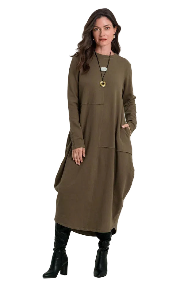 A woman modeling a long-sleeved, knee-length 100% cotton olive green dress with large front patch pockets, styled with black knee-high boots against a plain white studio background.