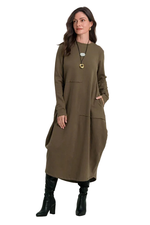 A woman modeling a long-sleeved, knee-length 100% cotton olive green dress with front patch pockets, styled with black ankle boots and a gold pendant necklace, set against a plain white studio background.