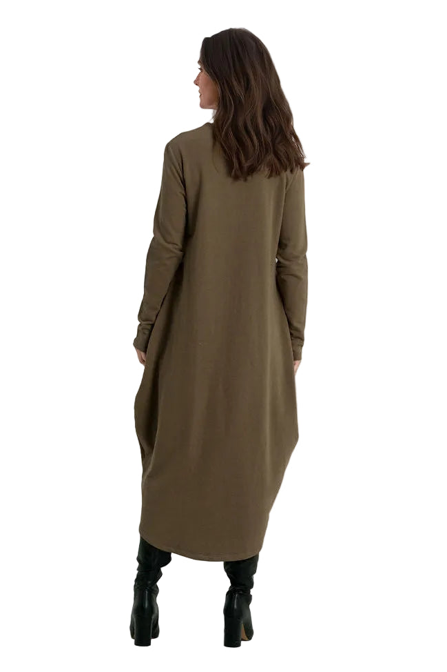 A woman viewed from behind wearing a long, 100% cotton loose-fitting olive brown dress or tunic with black knee-high boots, standing against a plain white studio background.