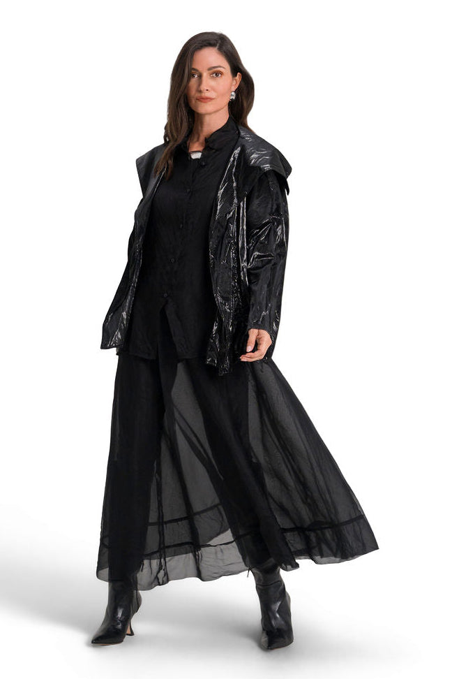 A woman modeling a black outfit consisting of a shiny black jacket, a black long-sleeve top, a sheer black midi skirt, and black pointed boots, photographed against a solid white studio background.