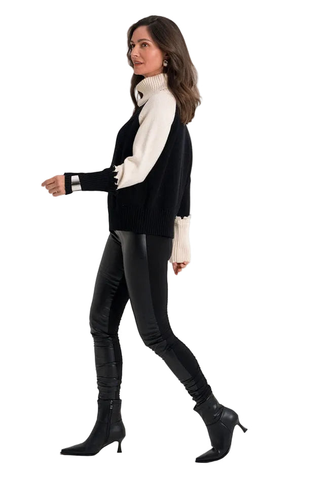 A woman modeling a black sleeveless vest over a white long-sleeve turtleneck, paired with black faux-leather leggings and black heeled ankle boots, captured in profile against a solid white studio background.