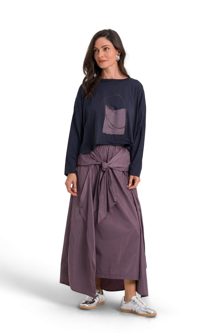 A woman modeling a 100% pima cotton dark navy long-sleeve top with a circular chest graphic and a dusty purple wrap-style midi skirt tied at the waist, paired with white sneakers, against a plain white studio background.