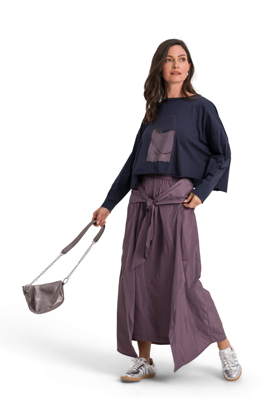 A woman modeling a 100% pima cotton dark navy blue cropped long-sleeve top with a large front patch pocket, paired with a high-waisted, wide-leg purple skirt and carrying a small gray shoulder bag, set against a plain white studio background.