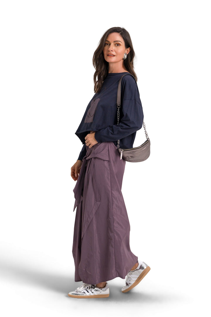 A woman modeling a 100% pima cotton dark navy blue long-sleeve top and matching wide-leg pleated skirt in a dusty purple shade, styled with white sneakers and a small shoulder bag against a white background.