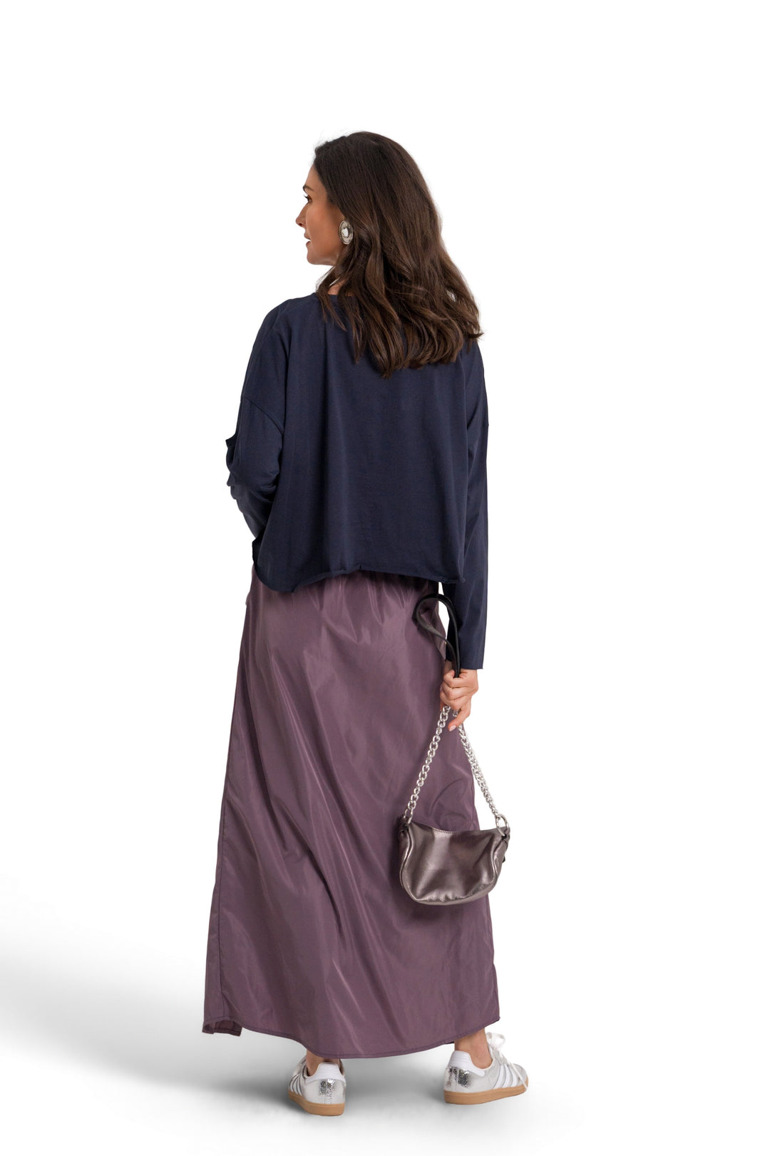 A woman is shown from behind wearing a 100% pima cotton navy blue long-sleeve top, a dusty purple maxi skirt, and holding a metallic silver shoulder bag against a solid white background.