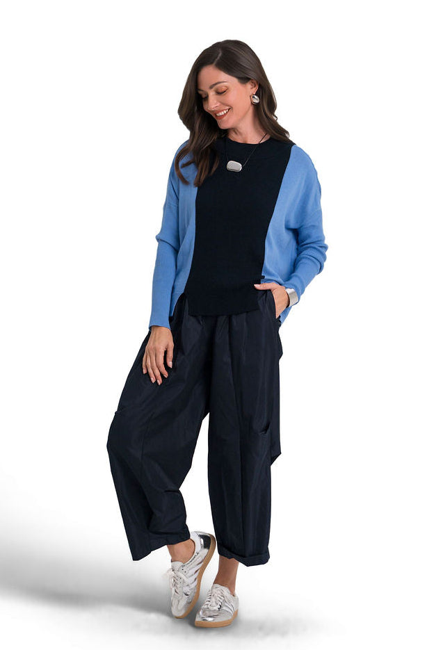 A woman modeling a two-tone pima cotton sweater with a blue body and dark navy sleeves, worn over a black top and navy wide-leg pants, on a white studio background.