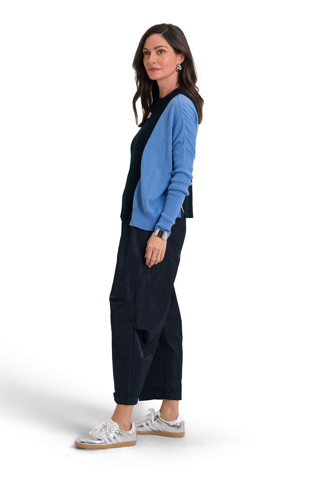 A woman stands in profile wearing a light blue long-sleeve pima cotton sweatshirt layered over a dark navy vest, paired with dark navy wide-leg cropped pants and white sneakers, against a plain white studio background.