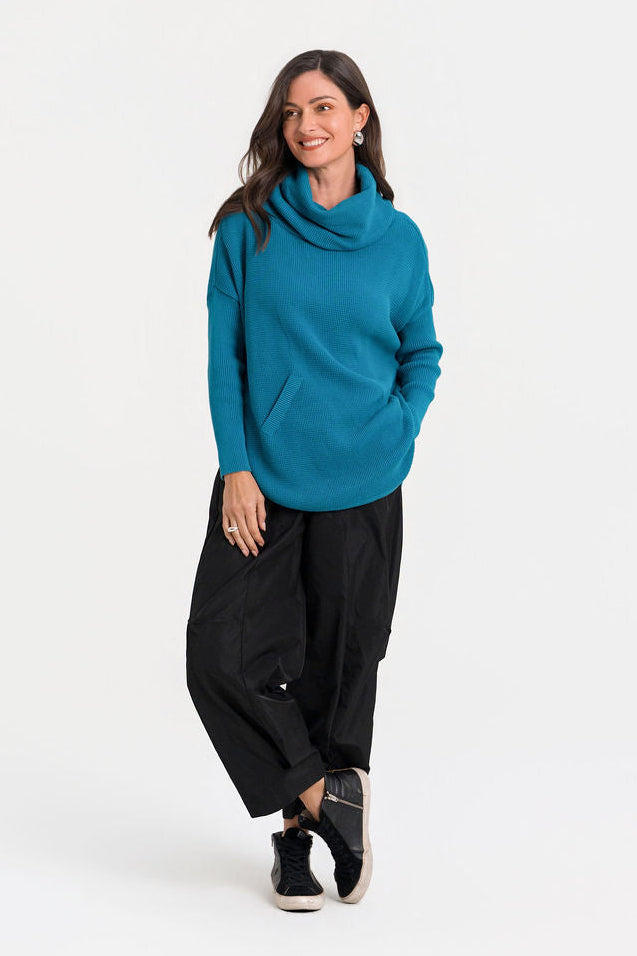 A woman modeling a teal blue cowl-neck knit pima cotton sweater and black wide-leg pants against a plain white studio background.