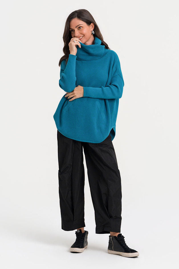 A woman modeling a teal blue pima cotton cowl-neck sweater, black wide-leg cropped pants, and black sneakers against a plain white studio background.