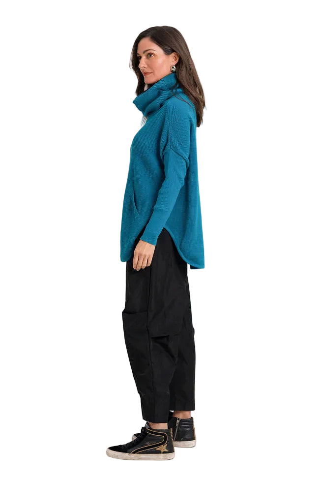 A woman shown in profile wearing a teal turtleneck sweater, black cargo pants, and black sneakers against a plain white background.
