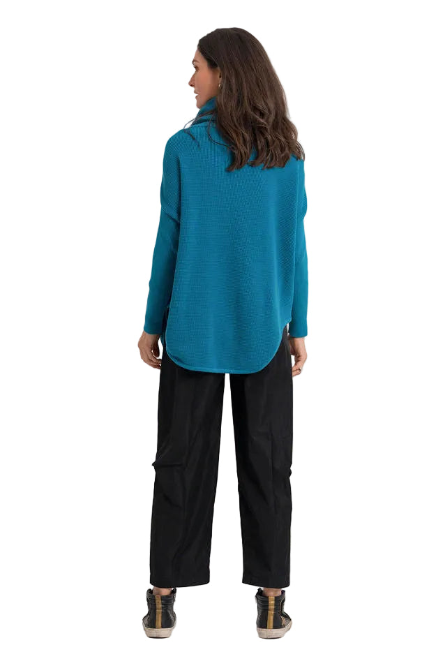 A woman shown from the back wearing a teal long-sleeve mock-neck top, black wide-leg pants, and black sneakers with gold details, set against a plain white background.