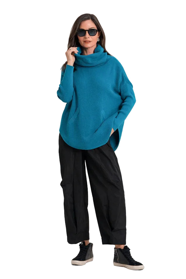 A woman modeling a teal blue oversized pima cotton knit cowl-neck sweater, paired with black wide-leg pants and black sneakers, against a plain white background.