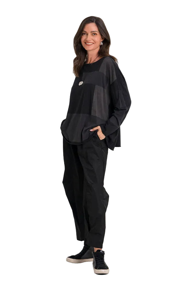 A woman modeling a 100% pima cotton black asymmetrical layered top and matching black wide-leg trousers, paired with black sneakers, against a seamless white studio background.
