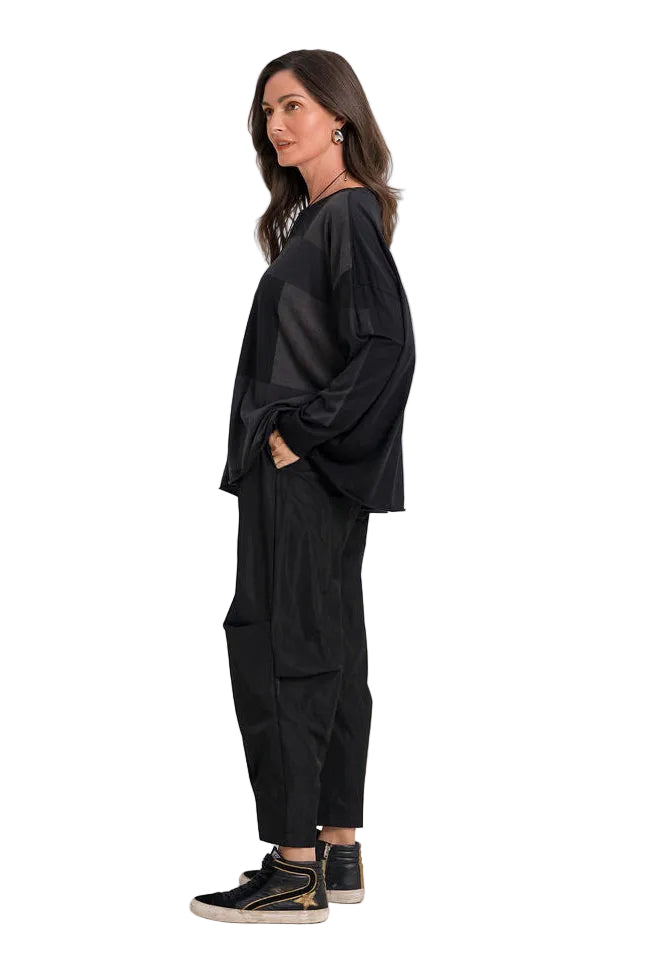 A woman stands in profile wearing a 100% pima cotton black long-sleeve top and black wide-leg pants, paired with black and gold sneakers, against a plain white studio background.