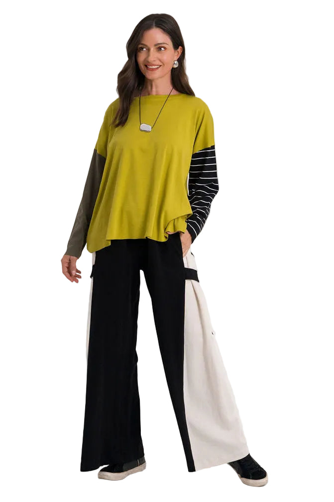 A woman modeling a color-blocked long-sleeve top with a mustard yellow body, black and white striped sleeves, and 100% cotton black wide-leg pants, standing against a plain white studio background.