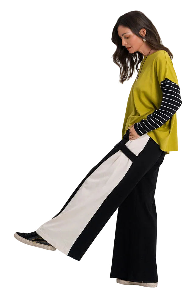 A woman modeling a color-blocked outfit consisting of a mustard yellow top with black and white striped sleeves, paired with 100% cotton wide-leg black pants featuring a large white side panel, set against a plain white studio background.