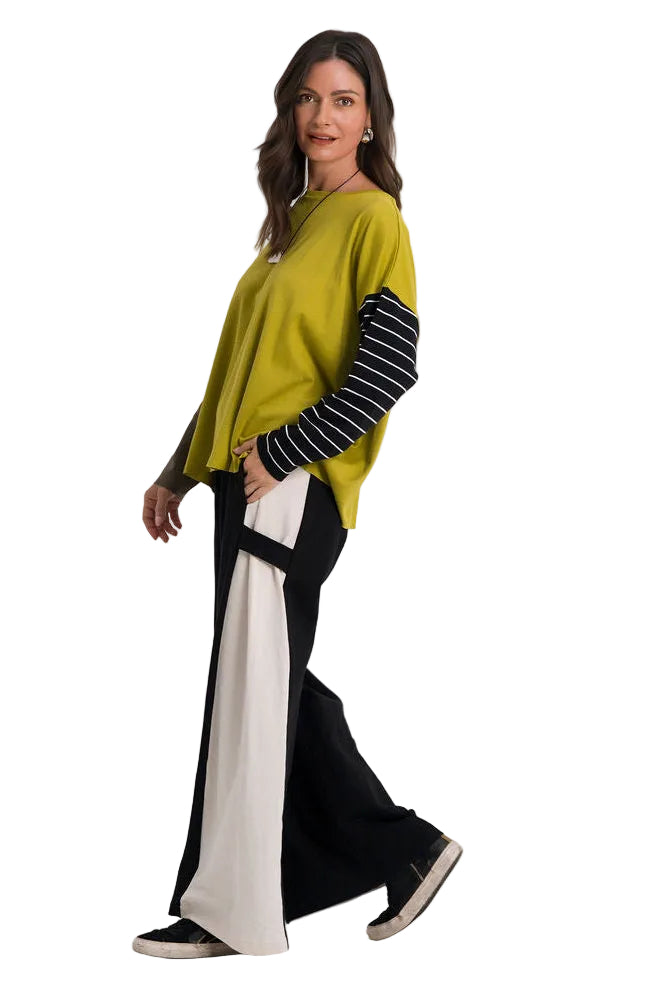 A woman modeling a color-blocked knit sweater with a mustard yellow body, black and white striped sleeves, and 100% cotton black wide-leg pants, walking against a plain white studio background.