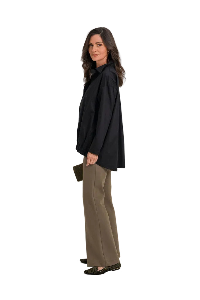 A woman modeling a 100% cotton black oversized long-sleeve blouse with wide-leg taupe-brown trousers, standing in profile against a clean off-white studio background.