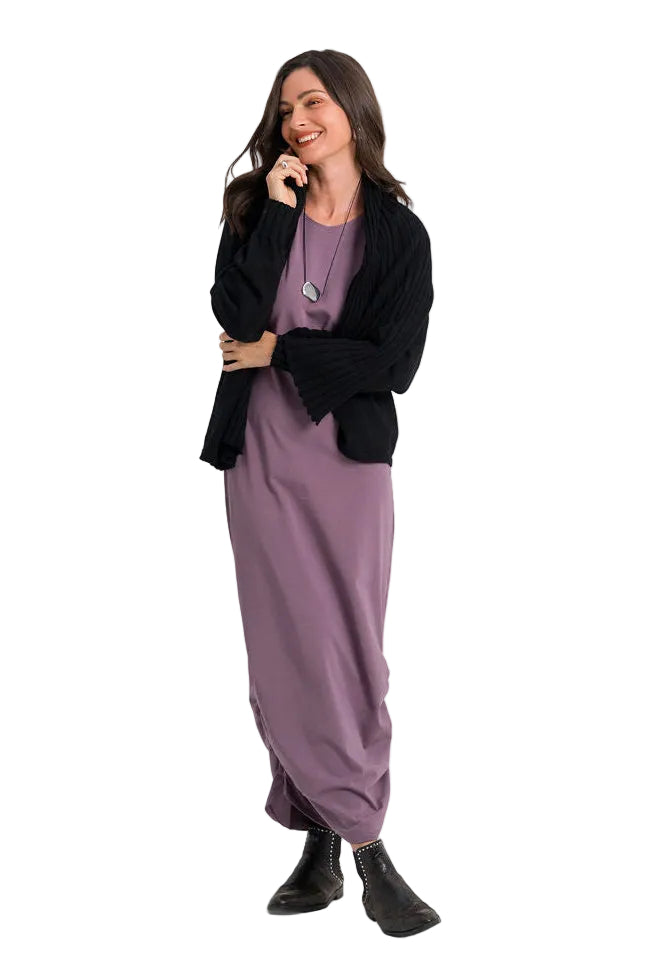 A woman modeling a loose-fitting mauve-purple maxi dress with a black cardigan, silver pendant necklace, and black ankle boots, standing against a plain white studio background.