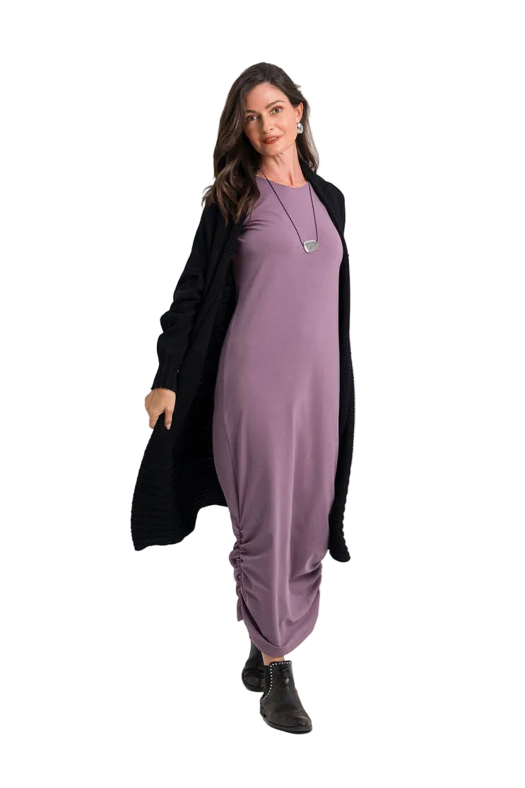 A woman modeling a long-sleeved mauve-purple maxi dress with gathered draping at the hem, styled with a black open-front cardigan, a silver pendant necklace, and black ankle boots against a plain white studio background.