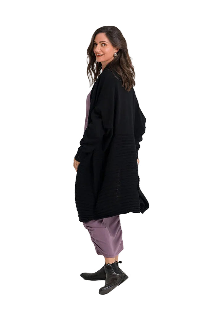 A woman modeling a long black open-front cardigan over a mauve dress, paired with black ankle boots, photographed against a plain white studio background.