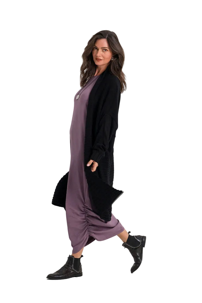 A woman modeling a long-sleeved black cardigan over a mauve-purple maxi dress with gathered hems, paired with black ankle boots, against a plain white studio background.