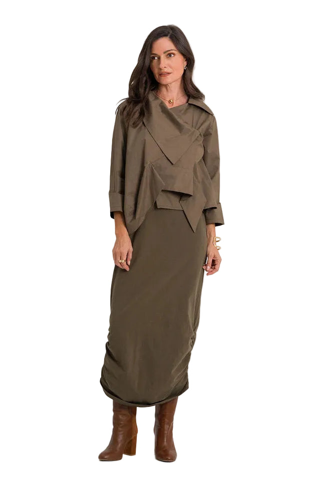 A woman modeling a matching 100% cotton two-piece outfit consisting of a draped, wrap-style long-sleeve top and a matching high-waisted midi skirt in olive green, paired with brown leather ankle boots, against a plain white studio background.