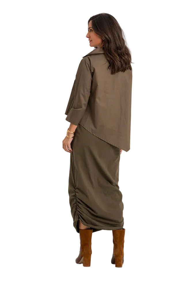 A woman shown from the back wearing an 100% cotton olive brown draped shirt dress with wide three-quarter sleeves and gathered ruching at the hem, paired with brown boots, against a plain white studio background.
