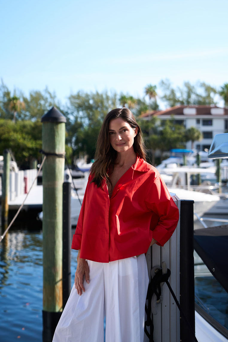 Woman in a red cotton shirt and white cotton pants standing by a dock with boats in the background
