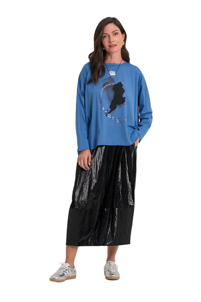 A woman modeling a 100% pima cotton blue crewneck sweatshirt with a dark graphic print, paired with a shiny black midi skirt and gray sneakers, against a plain white studio background.