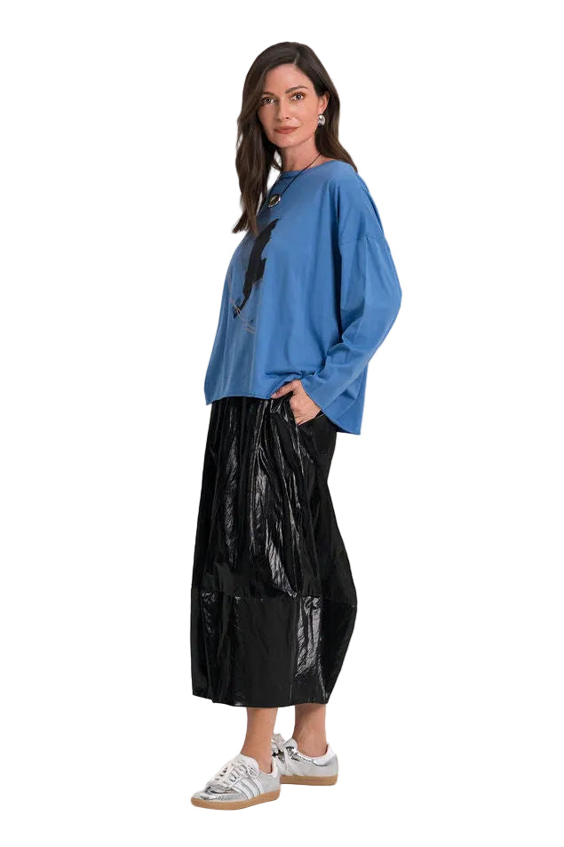 A woman modeling a 100% pima cotton blue long-sleeve top and a shiny black pleated skirt, paired with white sneakers, against a plain white studio background.