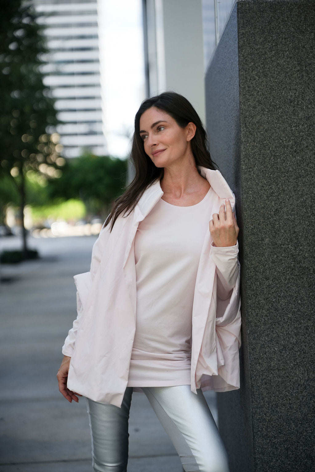 Woman in a light pink chic cape and blush shirt leaning against a building.