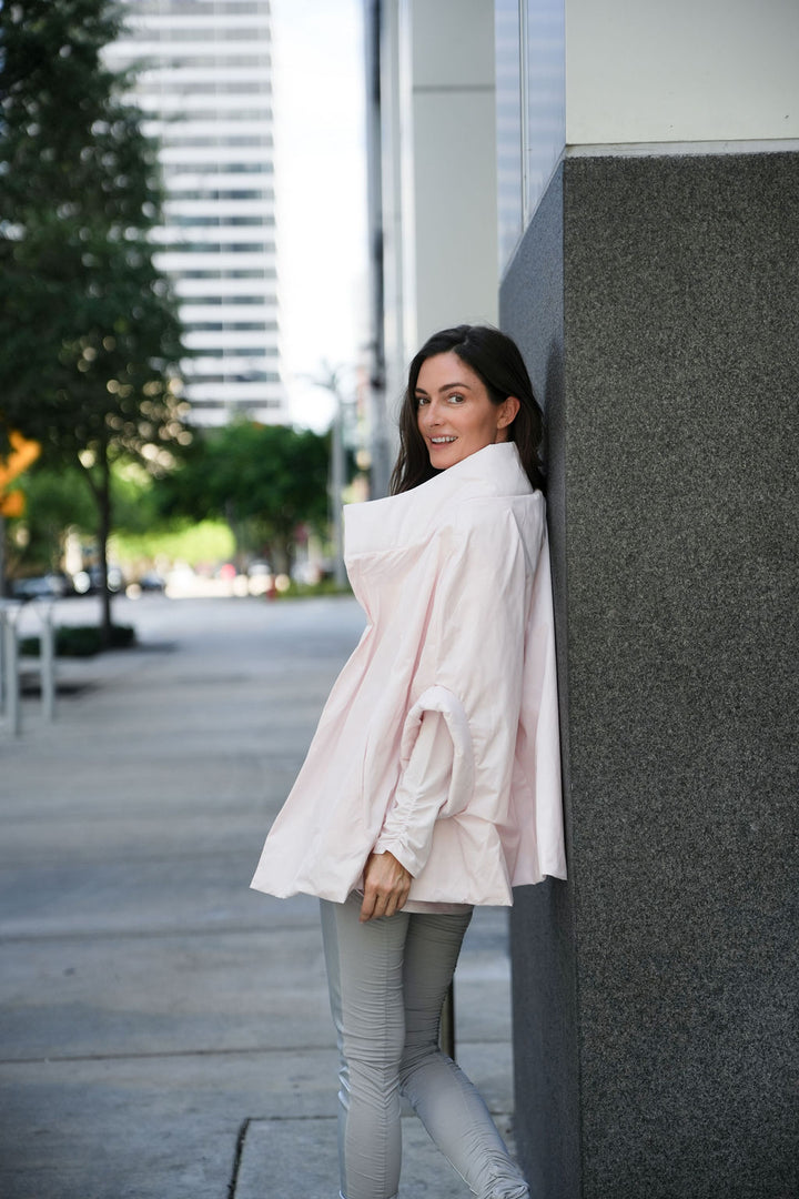 Woman in a light pink blush chic cape standing against a building wall on a city street.
