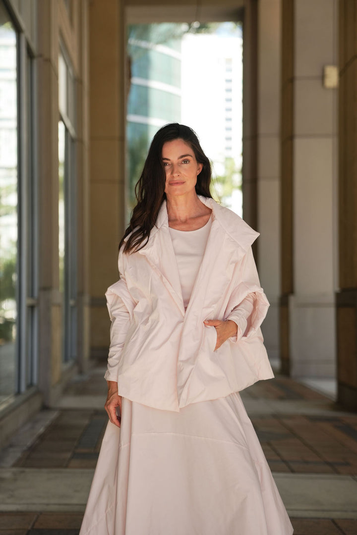 Woman in a light pink blush outfit standing in a sunlit hallway.