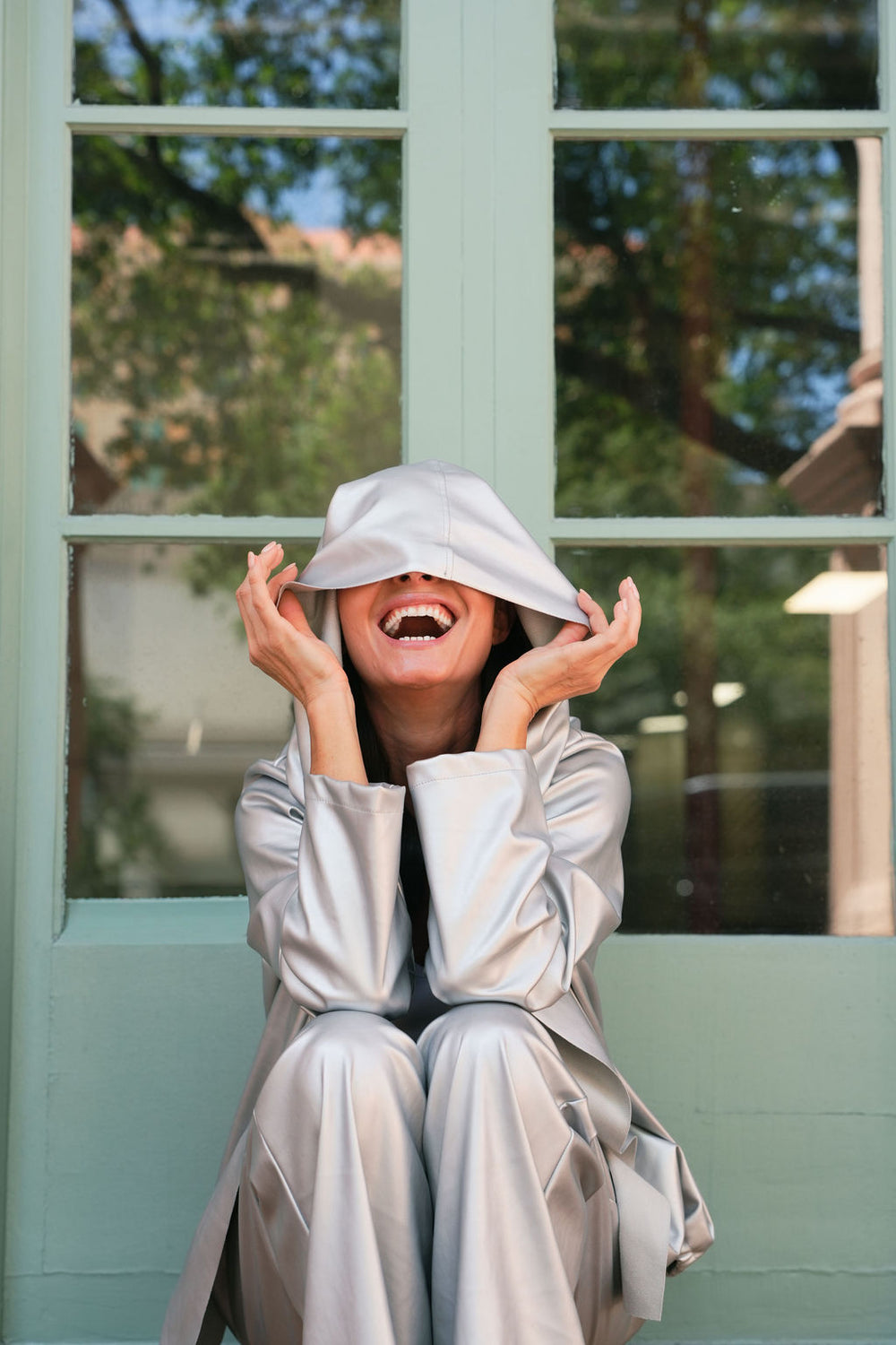 Smiling woman wearing a silver vegan leather hooded outfit sitting in front of a window with a view of trees.