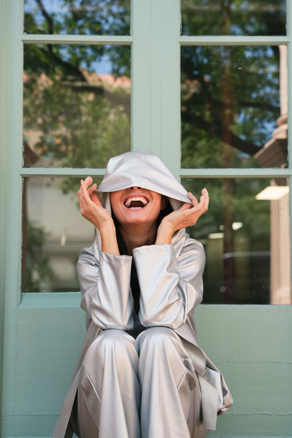 Smiling woman wearing a silver vegan leather hooded outfit sitting in front of a window with a view of trees.