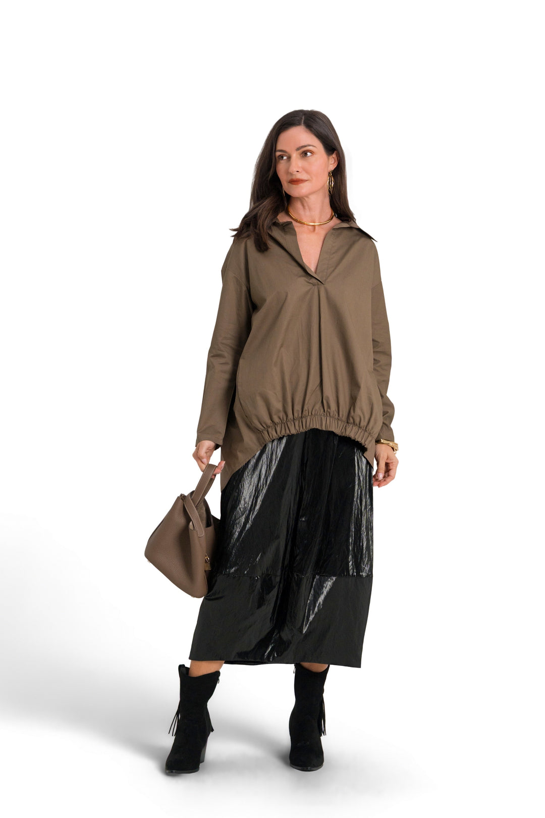 A woman stands against a white studio background wearing a 100% cotton brownish olive long-sleeve top, a shiny black skirt, black ankle boots, and holding a brown handbag.