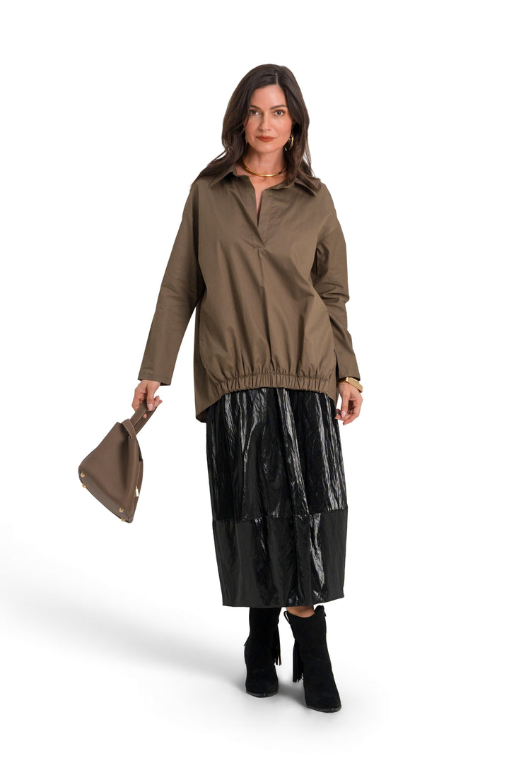 A woman modeling a 100% cotton brownish-olive long-sleeve jacket with a black pleated skirt and black boots, holding a matching brown clutch bag against a plain white background.