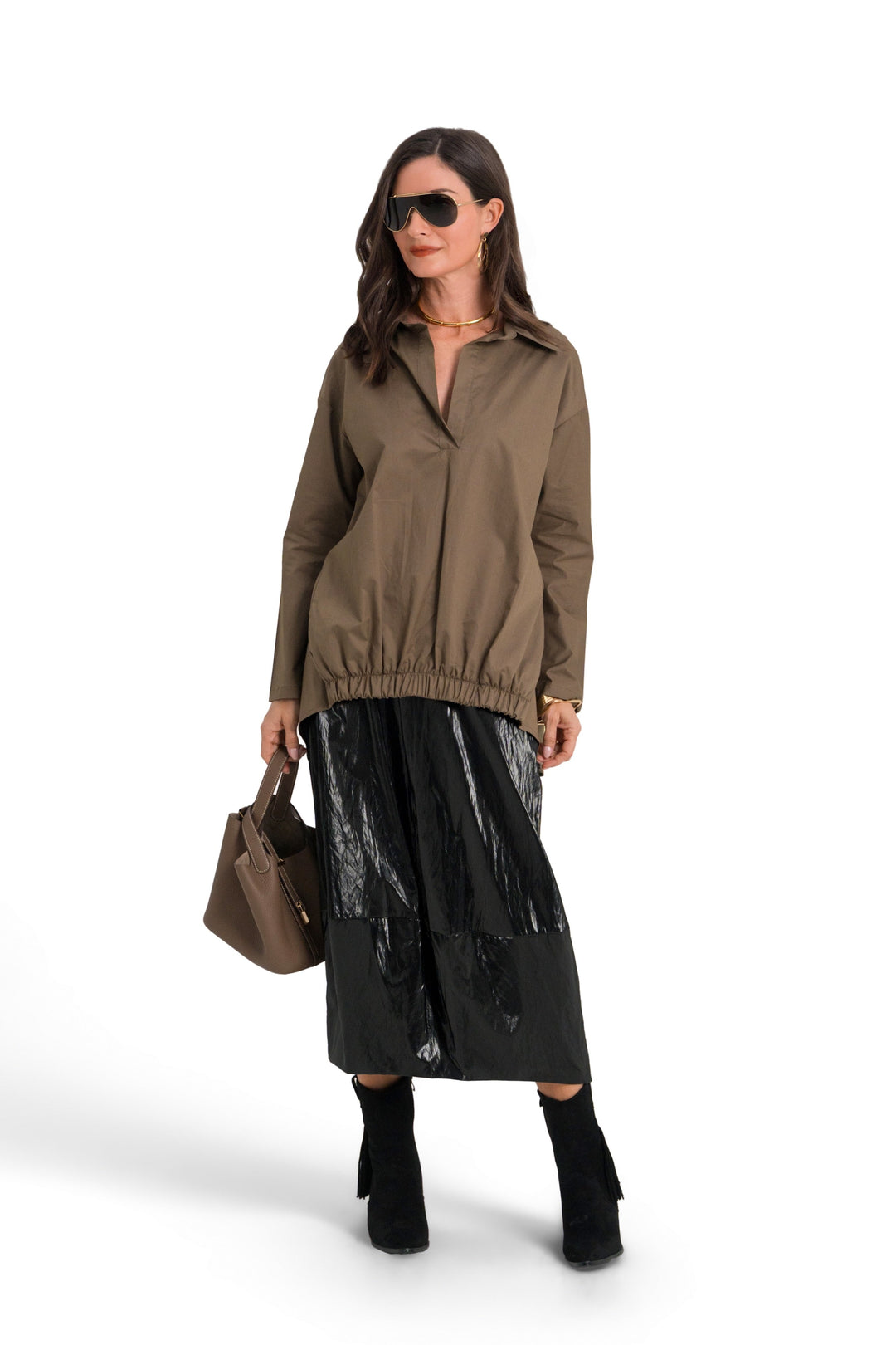 A model stands against a white studio background wearing a 100% cotton  brownish olive zip-up jacket, a shiny black midi skirt, black knee-high socks, and sunglasses, while holding a brown handbag.