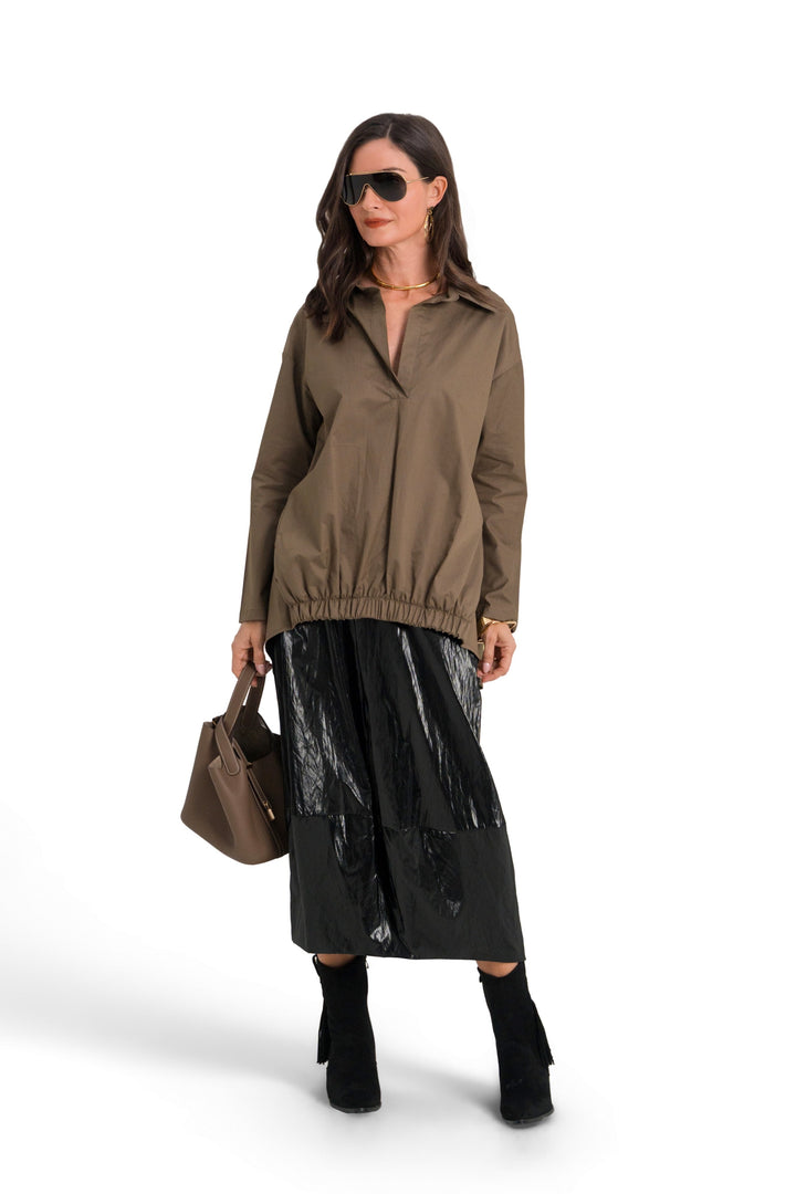 A model stands against a white studio background wearing a 100% cotton  brownish olive zip-up jacket, a shiny black midi skirt, black knee-high socks, and sunglasses, while holding a brown handbag.
