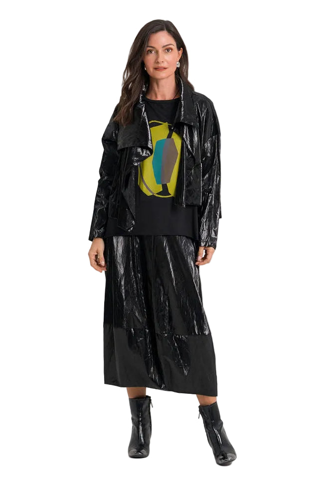 A model stands against a plain white studio background wearing a glossy black outfit consisting of a shiny jacket, a black midi skirt, a 100% pima cotton black top with a colorful graphic print, and black ankle boots.
