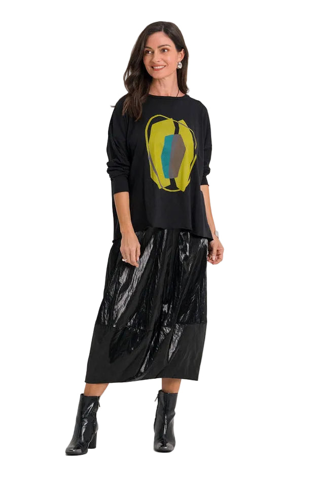 A woman modeling a 100% pima cotton black long-sleeve top with a yellow and blue graphic print, paired with a black pleated skirt and black ankle boots, against a plain white studio background.