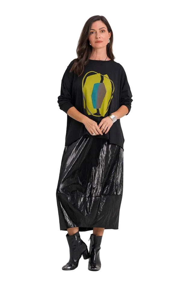 A woman modeling a 100% pima cotton black long-sleeve top with a yellow and blue graphic print, paired with a shiny black midi skirt and black ankle boots, against a plain white studio background.